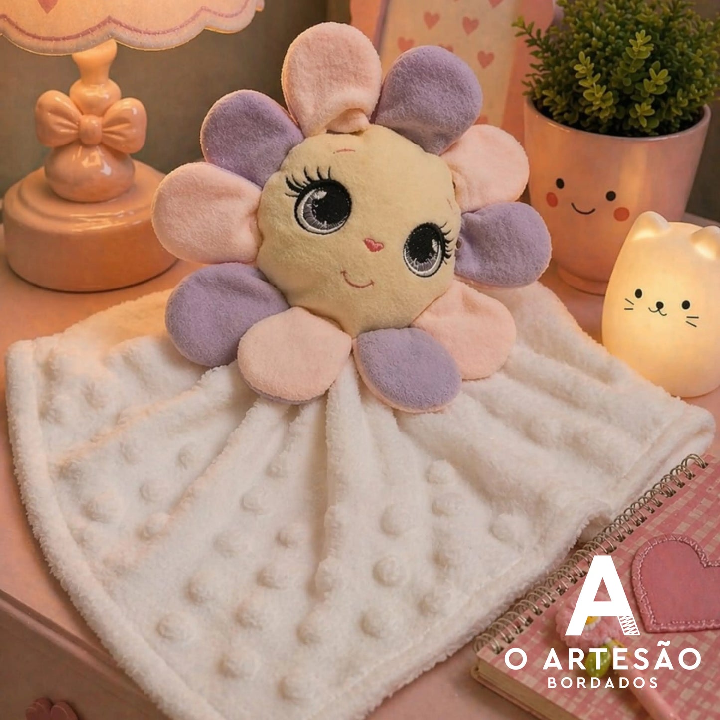 Naninha florzinha com pétalas lilás e rosa e rosto bordado com olhos grandes, apoiada sobre tecido branco acolchoado em cômoda cor-de-rosa
